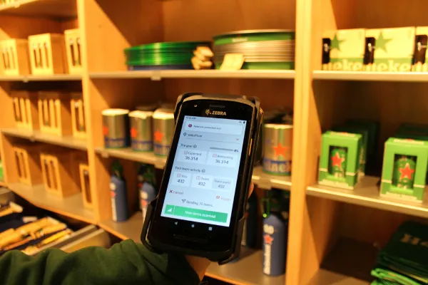 A handheld Zebra mobile computer displaying real-time RFID inventory data in front of Heineken merchandise shelves at the Heineken Experience store.