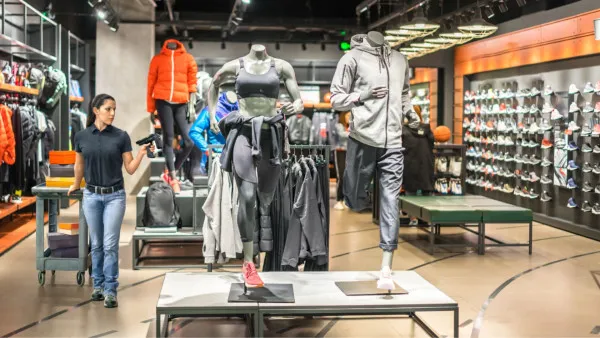 Retail store associate using a handheld RFID scanner for real-time inventory management in a sportswear and fashion retail environment, standing near a product display cart.